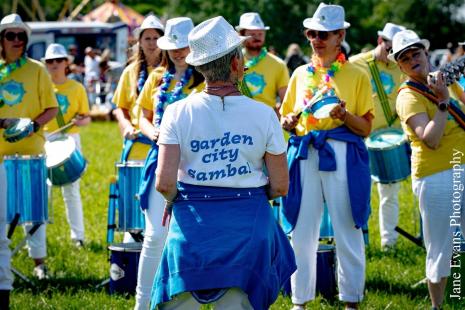 Group of people wearing yellow tops, white bottoms and blue jumpers tied around their waist playing drums outside on grass